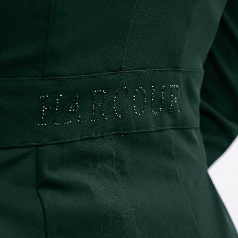 Seitliches Detailbild der Rückseite des Harcour Turnierjackets Oria in Jungle, einem Grünton. Im Bereich der Taille betont ein angedeuteter Bund den femininen Schnitt der Turnierjacke für Damen. Ein funkelnder Harcour-Logo-Schriftzug aus großen, grünen Strasssteinen ziert diesen Taillenbund. Dieses wunderschöne Glitzerdetail wertet die dezente Rückansicht auf und macht das Reitsakko für Markenfans zu einem echten Designerstück, das die weibliche Silhouette im Turniersport elegant in Szene setzt.
