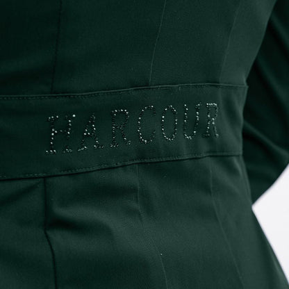 Seitliches Detailbild der Rückseite des Harcour Turnierjackets Oria in Jungle, einem Grünton. Im Bereich der Taille betont ein angedeuteter Bund den femininen Schnitt der Turnierjacke für Damen. Ein funkelnder Harcour-Logo-Schriftzug aus großen, grünen Strasssteinen ziert diesen Taillenbund. Dieses wunderschöne Glitzerdetail wertet die dezente Rückansicht auf und macht das Reitsakko für Markenfans zu einem echten Designerstück, das die weibliche Silhouette im Turniersport elegant in Szene setzt.