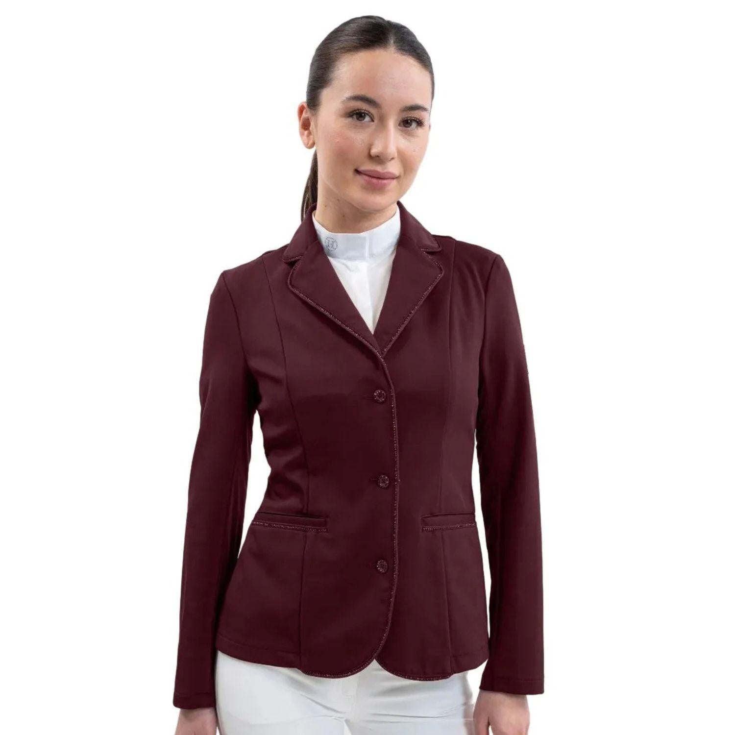 Eine Turnierreiterin präsentiert das feminine Harcour Damen Turnierjacket Oria in Aubergine, einem luxuriösen, dunkelroten Farbton, der Violett- und Brauntöne elegant vereint. Das figurbetonte Reitsakko mit hochelastischem Harcour-Riders-Stretch wird mit weißem Harcour Turniershirt und weißer Turnierreithose kombiniert. Die rahmende Glitzerlitze von Kragen bis Saum, betonte vordere Einschubtaschen sowie mit Kristallsteinen verzierte Knöpfe an Ärmeln und Verschluss machen die Reitjacke zum Blickfang.