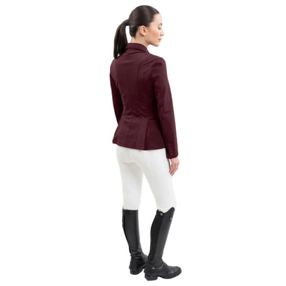 Rückansicht einer Reiterin im Harcour Damen Turnierjacket Oria. Die luxuriöse Farbe Aubergine, ein dunkler Rotton mit kühlen Violett- und warmen Brauntönen, harmoniert perfekt mit weißer Turnierkleidung. An der Taille funkelt der edle Harcour Glitzerschriftzug. Das elegante Schößchen, oft auch Reitrock genannt, bietet dank zwei seitlichen Schlitzen höchste Flexibilität und Bewegungsfreiheit im Sattel. Ein exklusives Reitsakko, das im Turniersport durch edles Design perfekt in Szene gesetzt wird.