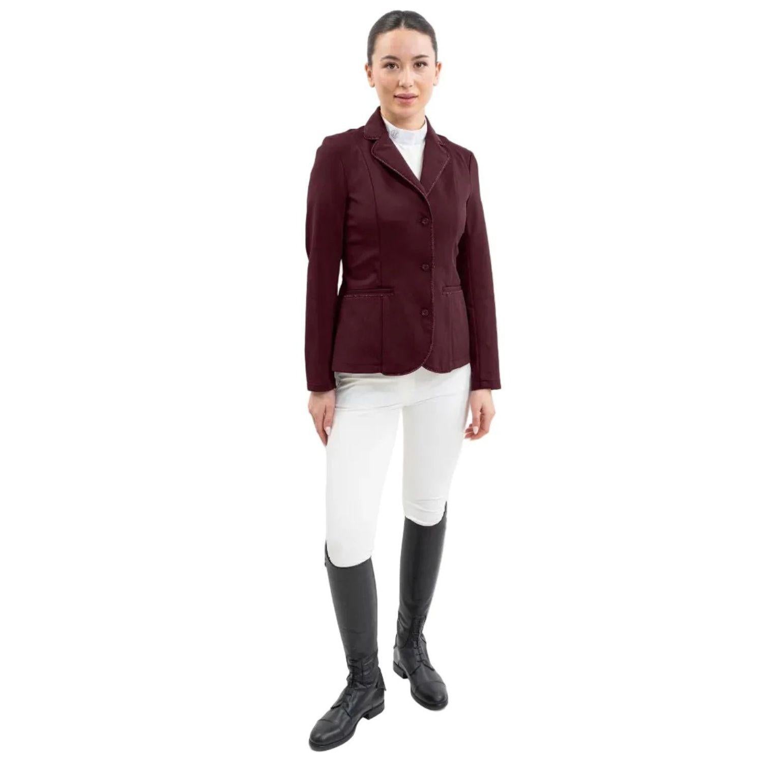 Eine Turnierreiterin präsentiert ein komplettes Harcour Turnieroutfit. Sie trägt das Damen Turnierjacket Oria in Aubergine, kombiniert mit weißer Turnierreithose und dem Harcour Turniershirt Shine mit weißem Latz. Dieses edle Reitsakko setzt optische Highlights und bietet maximale Sicherheit: Es ist kompatibel mit dem Harcour Ouraye HIT AIR Airbag und allen Airbagwesten, die unter der Turnierkleidung getragen werden. Ein elegantes und sicheres Reitoutfit für den anspruchsvollen Turniersport.