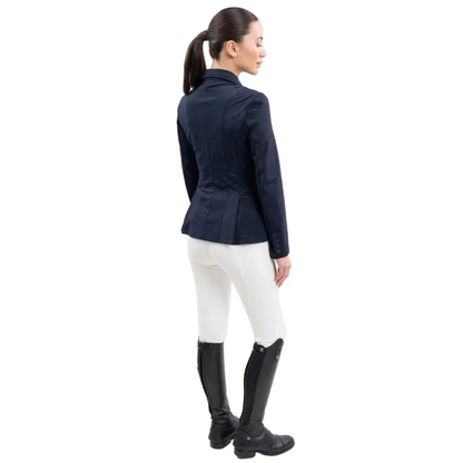 Rückansicht einer Reiterin im Harcour Damen Turnierjacket Oria. Die luxuriöse Farbe Navy, ein dunkler Blauton auch Marine genannt, harmoniert perfekt mit weißer Turnierkleidung. An der Taille funkelt der edle Harcour Glitzerschriftzug. Das elegante Schößchen, oft auch Reitrock genannt, bietet dank zwei seitlichen Schlitzen höchste Flexibilität und Bewegungsfreiheit im Sattel. Ein exklusives Reitsakko, das im Turniersport durch edles Design perfekt in Szene gesetzt wird.