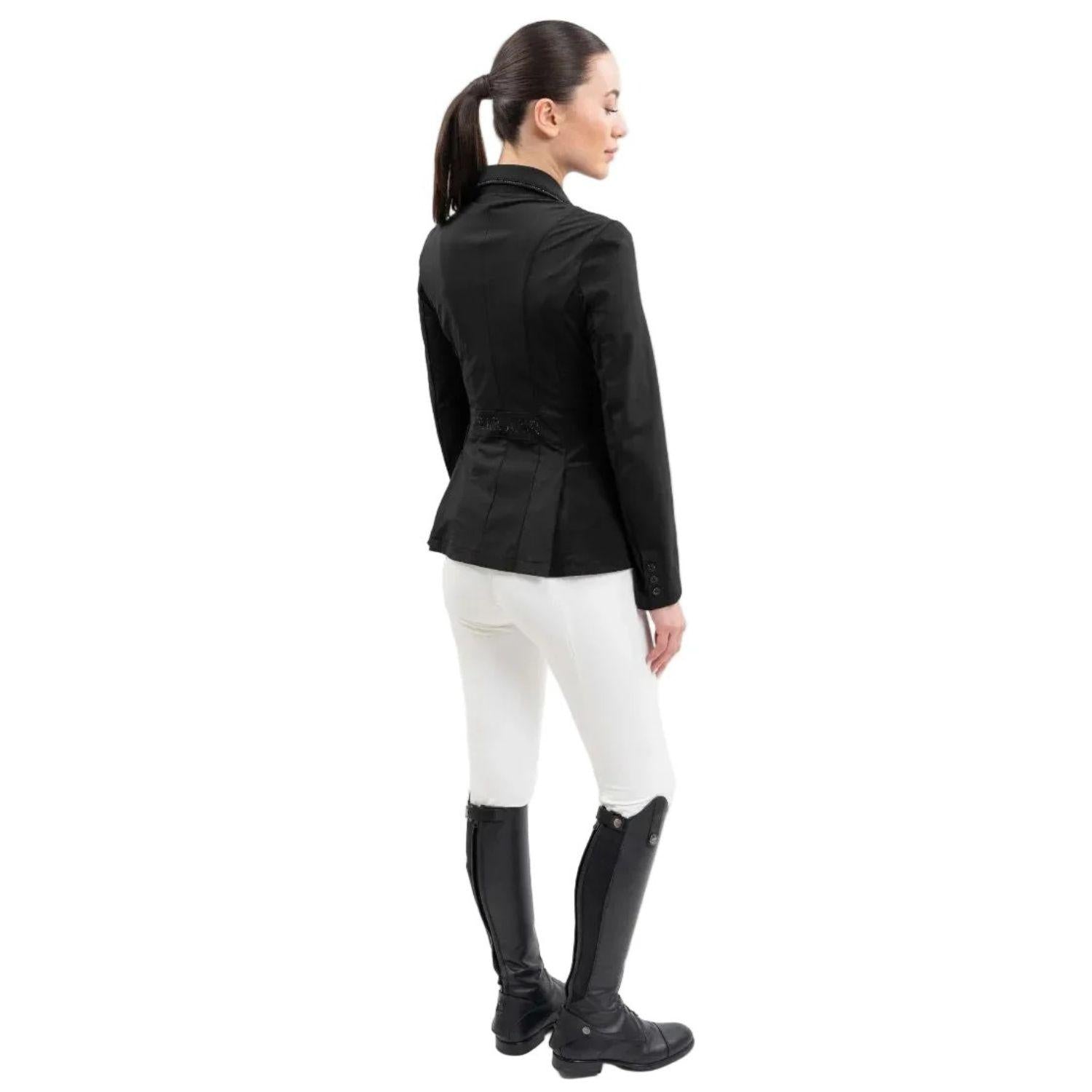 Rückansicht einer Reiterin im Harcour Damen Turnierjacket Oria. Die luxuriöse Farbe Schwarz harmoniert perfekt mit weißer Turnierkleidung. An der Taille funkelt der edle Harcour Glitzerschriftzug. Das elegante Schößchen, oft auch Reitrock genannt, bietet dank zwei seitlicher Schlitze höchste Flexibilität und Bewegungsfreiheit im Sattel. Ein exklusives Reitsakko, das im Turniersport durch edles Design perfekt in Szene gesetzt wird.