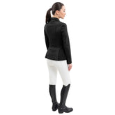 Rückansicht einer Reiterin im Harcour Damen Turnierjacket Oria. Die luxuriöse Farbe Schwarz harmoniert perfekt mit weißer Turnierkleidung. An der Taille funkelt der edle Harcour Glitzerschriftzug. Das elegante Schößchen, oft auch Reitrock genannt, bietet dank zwei seitlicher Schlitze höchste Flexibilität und Bewegungsfreiheit im Sattel. Ein exklusives Reitsakko, das im Turniersport durch edles Design perfekt in Szene gesetzt wird.