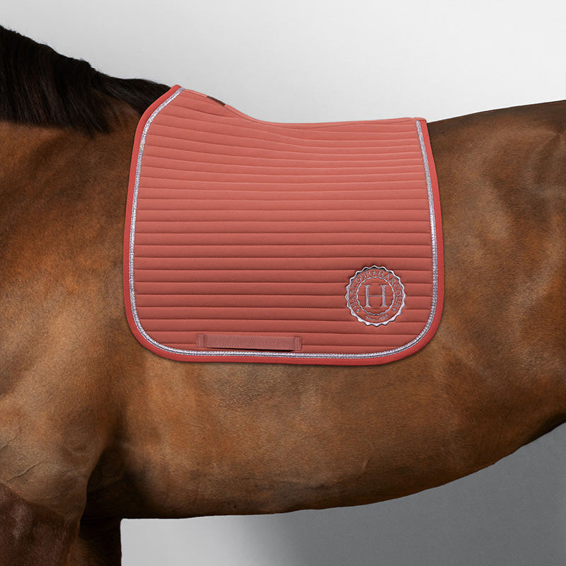 Harcour Schabracke Karembar in Aurora Pink am Pferd (Farbe: Braun, schwarzes Langhaar). Detailaufnahme der Dressurschabracke: anatomischer Schnitt mit großer Widerristfreiheit, elegante horizontale Steppung und die wunderschön glitzernde silberne flache Litze im Sonnenlicht. Das Harcour-Logo ist silber eingestickt. Die Farbharmonie ist perfekt: Das frische, helle Aurora Pink kontrastiert wunderschön mit dem satten Braun und dem schwarzen Langhaar des Pferdes. Eine hochwertige Sattelunterlage mit Glitzer.