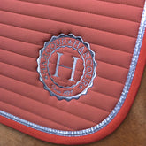 Gestochen scharfes Detailbild der linken unteren Seite der Harcour Schabracke Karembar in der Farbe Aurora Pink. Die Aufnahme zeigt die präzise horizontale Steppung, das farblich abgesetzte, dunklere rosa Einfassband und die flache, silberne glitzernde Litze. Diese funkelnde Litze passt perfekt zu dem edlen, runden, silber eingestickten Harcour Logo auf der Sattelunterlage. Ein echter stylischer Blickfang und Garant für einen perfekten Auftritt, egal ob im Dressursattel, Springsattel oder Ponysattel. 