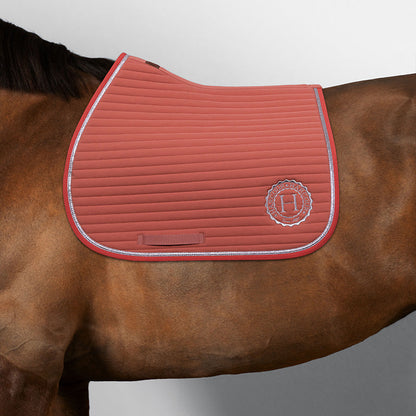 Harcour Schabracke Karembar Aurora Pink als Springschabracke auf einem braunen Pferd mit schwarzem Langhaar. Die anatomische Sattelunterlage passt perfekt unter alle modernen Springsättel und ist ideal für das Springtraining oder Ausritte. Hoher Schnitt am Widerrist, die stoßdämpfende horizontale Steppung und die wunderschöne silberne Glitzer-Litze. Dynamischer Farbkontrast: strahlendes Rosa hebt sich sportlich und frisch vom braunen Fell ab. Eine exzellente rosa Schabracke für den Springsport.