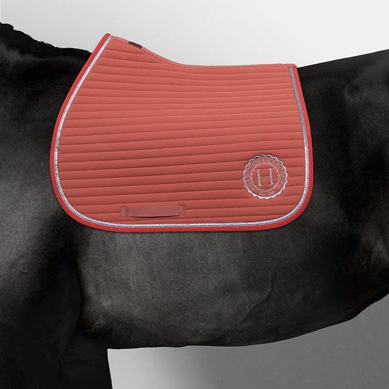 Harcour Springschabracke Karembar in Aurora Pink (Größe Warmblut) auf einem schwarzen Pferd (Rappe). Die Passform ist maßgeschneidert für gängige Springsättel und bietet höchsten Komfort beim Springtraining oder entspannten Ausritten. Die Aufnahme zeigt die optimale Widerristfreiheit, die klare Steppung und die funkelnde, silberne flache Litze. Spektakuläre Farbharmonie: Das leuchtende Rosa der Sattelunterlage bildet einen atemberaubenden, modernen Kontrast zum tiefschwarzen Fell. 