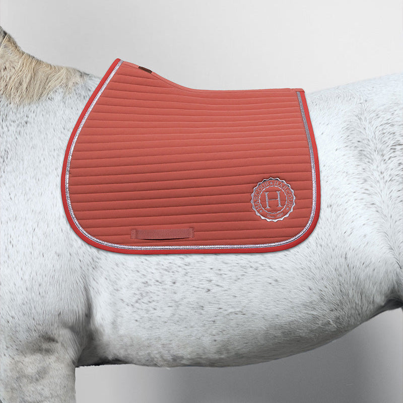 Harcour Sattelunterlage Karembar Aurora Pink auf einem weißen Pferd. Perfekt geschnitten für moderne Springsättel, schützt sie den Pferderücken zuverlässig bei täglichen Reiteinheiten, Ausritten und im Springtraining. Zu sehen ist die horizontale Steppung, das silberne Harcour-Logo und die elegante Glitzer-Litze. Die Farbharmonie ist absolut edel: Das zarte, frische Aurora Pink verschmilzt optisch mit dem hellen Fell des Schimmels. Ein absoluter Hingucker als rosa Schabracke mit Glitzer auf dem Springplatz.