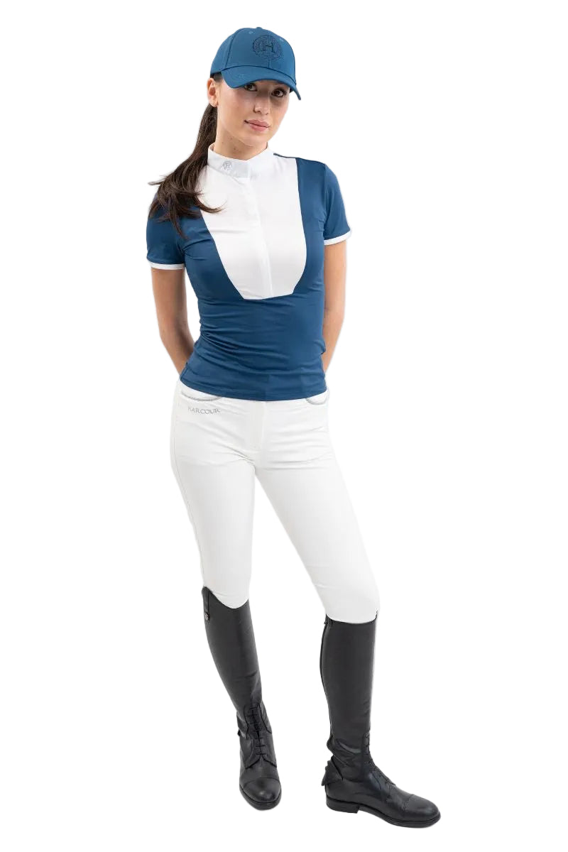 n einer eindrucksvollen Großaufnahme präsentiert eine Turnierreiterin ihr komplettes Harcour-Outfit, bestehend aus dem petrolfarbenen Polo-Kurzarm-Turniershirt Shine in Blue Lagoon, einer weißen Reithose und schwarzen Lederreitstiefeln, wobei das leichte, atmungsaktive Stretchmaterial mit dem klassischen weißen Latz feminine Modernität, höchsten Tragekomfort und sportliche Funktion vereint.