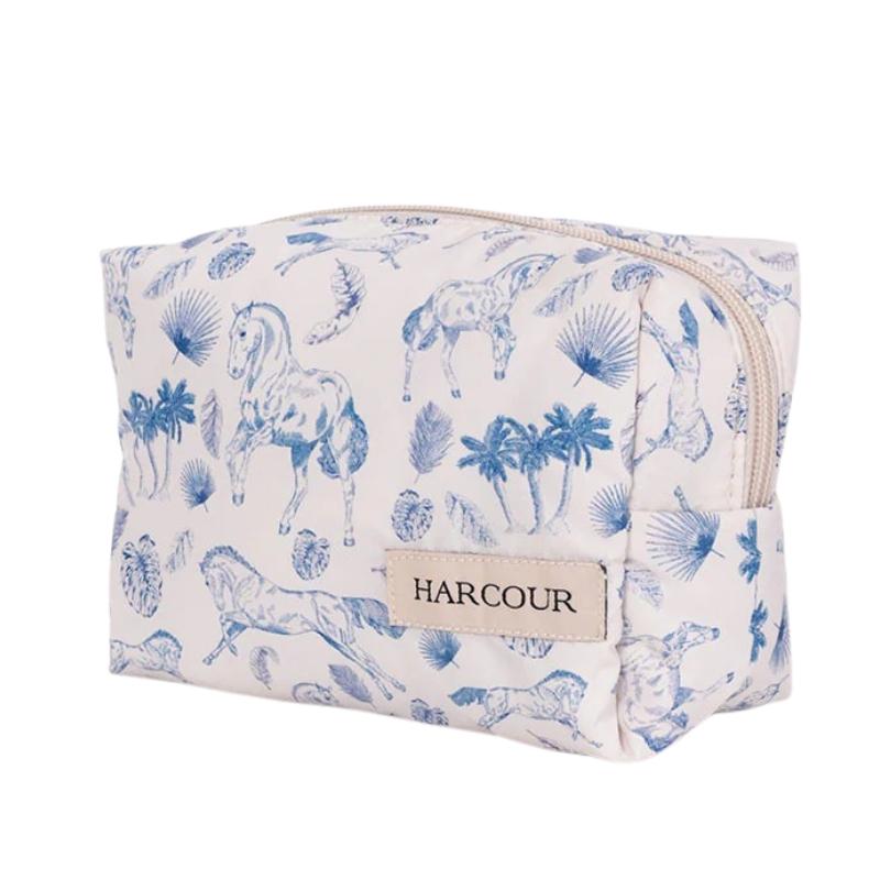 Die Harcour Kulturtasche Pam präsentiert sich in einer Front-Seitenansicht vor weißem Hintergrund mit geschlossenem, hellem Reißverschluss und zeigt ein edles, mediterranes Pferdeprint-Motiv in Blau mit Pferden in Bewegung, Palmen und Palmblättern. Das beige Harcour Logo-Patch ist rechts am unteren Teil der hellen Tasche angebracht, deren frisches Design den sportlichen Equestrian Lifestyle verkörpert.