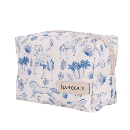 Die Harcour Kulturtasche Pam präsentiert sich in einer Front-Seitenansicht vor weißem Hintergrund mit geschlossenem, hellem Reißverschluss und zeigt ein edles, mediterranes Pferdeprint-Motiv in Blau mit Pferden in Bewegung, Palmen und Palmblättern. Das beige Harcour Logo-Patch ist rechts am unteren Teil der hellen Tasche angebracht, deren frisches Design den sportlichen Equestrian Lifestyle verkörpert.