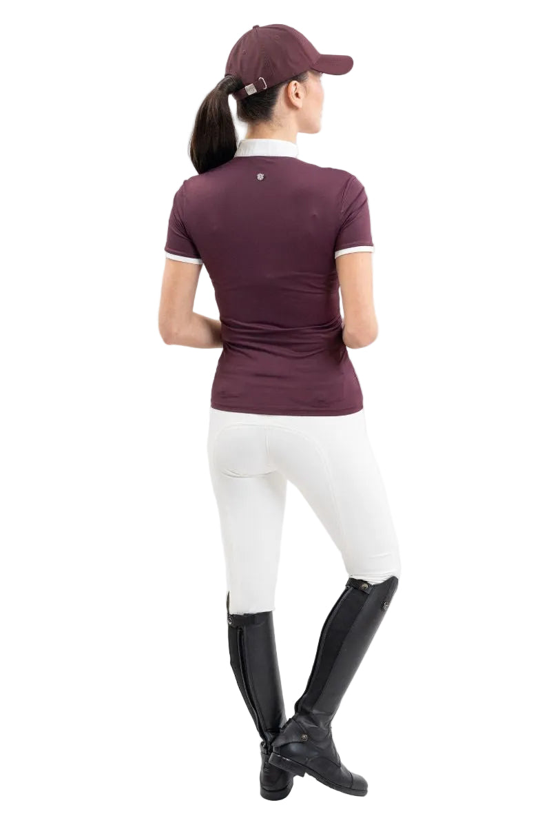 Die Rückansicht zeigt eine Turnierreiterin im eleganten Harcour Turniershirt Shine in der Farbe Aubergine kombiniert mit weißer Reithose und Reitstiefeln, wobei das französische Design mit besonderer Raffinesse und Funktionshighlights für absoluten Tragekomfort im Sattel sorgt.