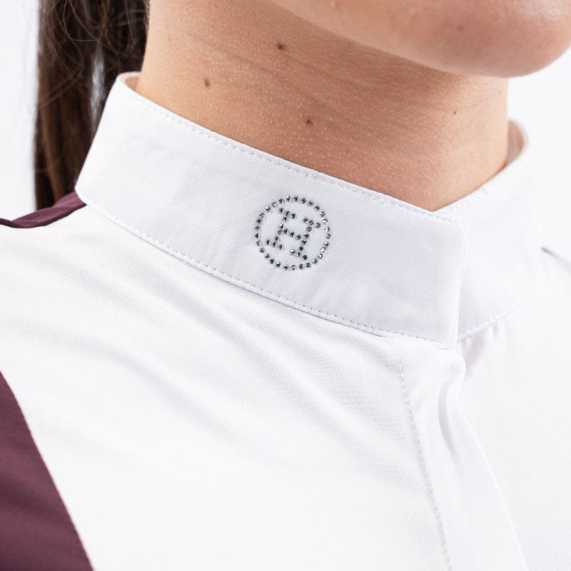 ine Nahaufnahme des eleganten weißen Stehkragens am Harcour Damen Turniershirt Shine Aubergine zeigt das funkelnde H-Logo aus hellen Kristallsteinen, welches unter dem Turnierjacket oder Dressurfrack für einen raffinierten Hauch von Glitzer sorgt und ein zusätzliches Plastron überflüssig macht. Verschluss durch einen Magneten und langen Reißverschluss zum An- und Ausziehen.