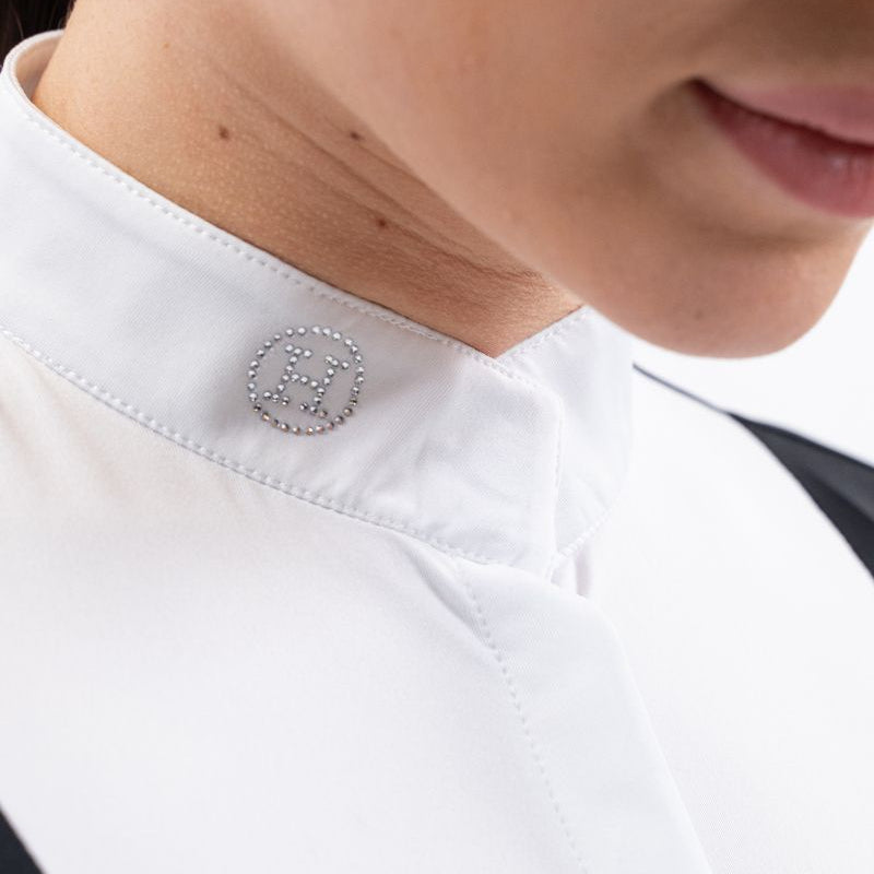 Eine Nahaufnahme des eleganten weißen Stehkragens am Harcour Damen Turniershirt Shine Black zeigt das funkelnde H-Logo aus hellen Kristallsteinen, welches unter dem Turnierjacket oder Dressurfrack für einen raffinierten Hauch von Glitzer sorgt und ein zusätzliches Plastron überflüssig macht. Verschluss durch einen Magneten und langen Reißverschluss zum An- und Ausziehen.