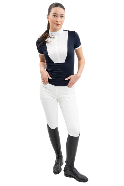 Eine Turnierreiterin im kompletten, eleganten Turnieroutfit trägt frontal das navyblaue Harcour Kurzarm-Turniershirt Shine mit edlem weißem Latz, passendem Stehkragen und kontrastfarbigen Ärmelabschlüssen, kombiniert mit einer weißen Harcour Reithose Jaltika und schwarzen Lederreitstiefeln für ein harmonisches Erscheinungsbild im Dressursport wie Springsport an warmen Sommertagen.