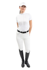 Eine Turnierreiterin im kompletten, eleganten Turnieroutfit trägt frontal das weiße Harcour Kurzarm-Turniershirt Shine mit Latz, Stehkragen mit Glitzer-Logo und Magnetverschluss, kombiniert mit einer weißen Harcour Reithose Jaltika und schwarzen Lederreitstiefeln für ein harmonisches Erscheinungsbild im Dressursport wie Springsport an warmen Sommertagen.