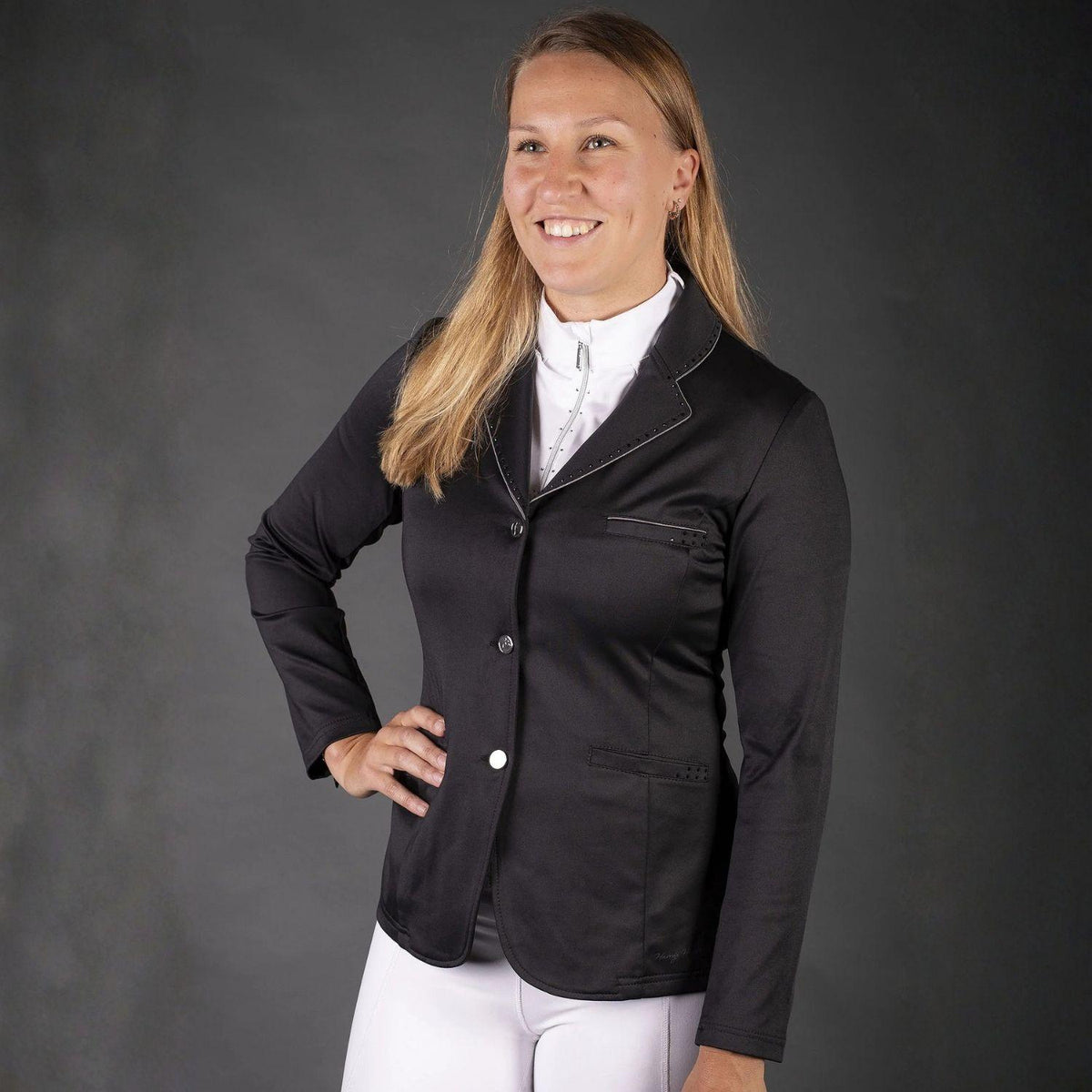 ine lächelnde Reiterin präsentiert das schwarze Harry’s Horse Turnierjacket Glitter Damen in getragener Ansicht vor grauem Hintergrund. Das taillierte Reitsakko aus elastischem 4-Wege-Stretch ist als komplettes Turnieroutfit mit einer weißen Turnierbluse und einer weißen Reithose kombiniert. Sichtbar sind die glitzernden Details (Biese &amp; Strass) an Kragen, Revers und Taschen sowie die silbernen Knöpfe. Sie hat die rechte Hand auf der Hüfte abgestützt, was die elegante, sportliche Passform betont.