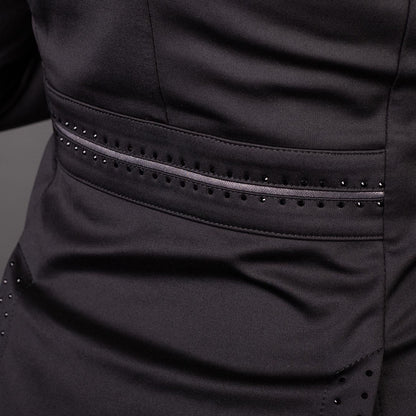 Makro-Detailaufnahme des Gürtelbands in Taillenhöhe auf der Rückseite des schwarzen Harry’s Horse Turnierjackets Glitter. Die taillierte Form des Reitsakkos wird durch eine mittig verlaufende graue Glitzerbiese betont, die oben und unten von präzise eingefassten, kleinen schwarzen Strasssteinen gerahmt ist. Am unteren Bildrand zeichnen sich zudem die dezent verzierten Ansätze der Gehschlitze des elastischen Stretch-Sakkos ab.