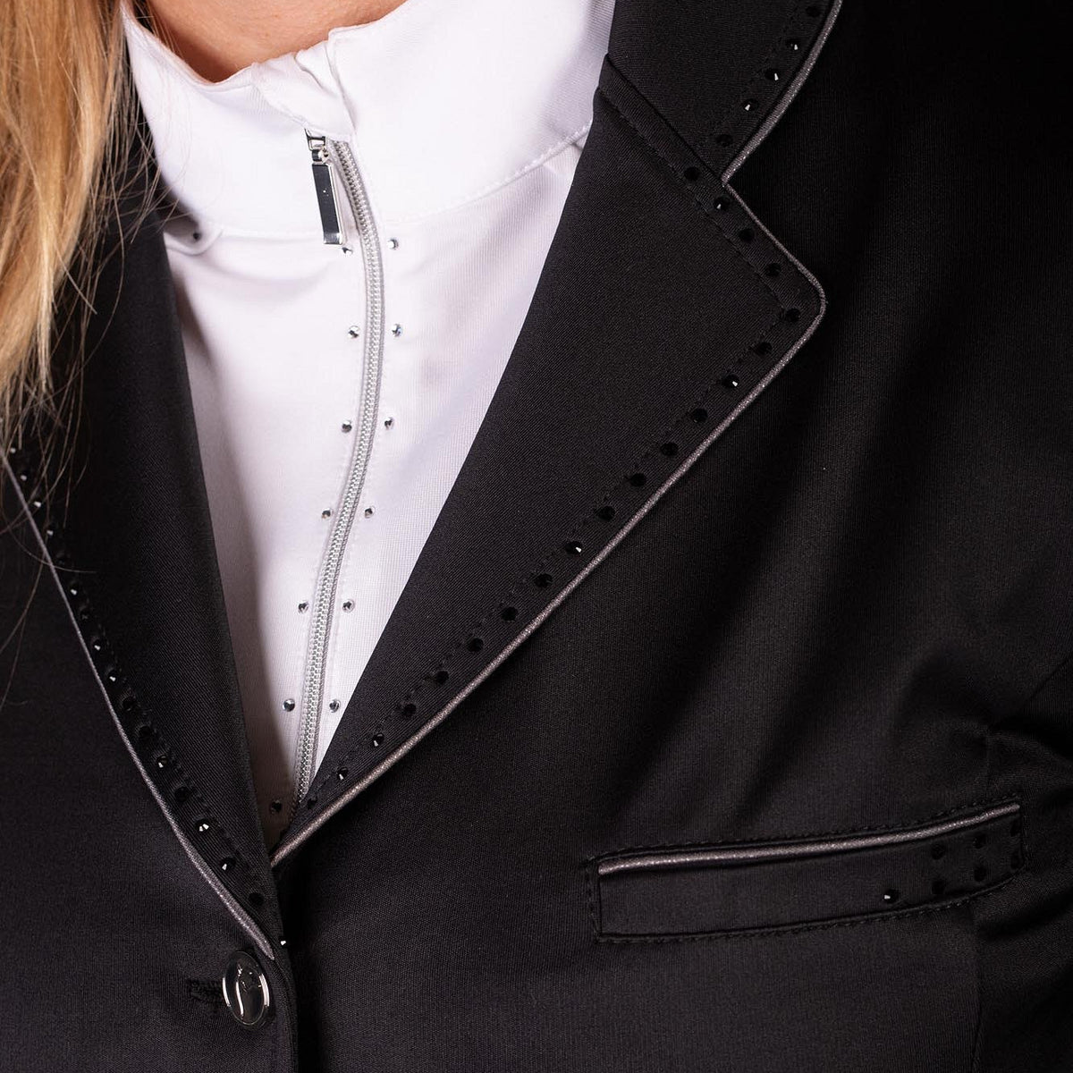 Nahaufnahme der edlen Verarbeitungsdetails des schwarzen Harry’s Horse Turnierjackets Glitter. Zu sehen sind der Kragen und das Revers des Reitsakkos, umrahmt von einer kontraststarken grauen Glitzerbiese und einer Reihe feiner schwarzer Strasssteine. Auch die Brusttasche auf der linken Seite greift das funkelnde Design mit Glitzerbiese und Strass-Akzenten auf. Darunter wird eine weiße Turnierbluse mit Stehkragen getragen, die den luxuriösen Gesamteindruck unterstreicht.