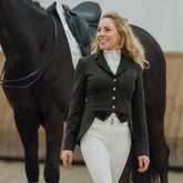 Eine Dressurreiterin im kompletten Turnieroutfit geht lachend neben ihrem Pferd und trägt das schwarze Harry’s Horse Turnierjacket Modell Montpellier im modernen Kurzfrack-Design. Die detailreichen Glitzer-Pailletten und Strasssteine am Kragen, an der Double-Closure-Leiste und den kurzen Dressurschößen kommen im natürlichen Tageslicht voll zur Geltung, betonen das edle Design und zeigen die makellose Silhouette des Kurzfracks.