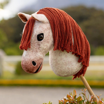 LeMieux Hobby Horse Poppy draußen vor einem Reitplatz. Das Rotschimmel-Fell zeigt leichte Rotverläufe im Kontrast zur roten Mähne und dem plüschigen Maul