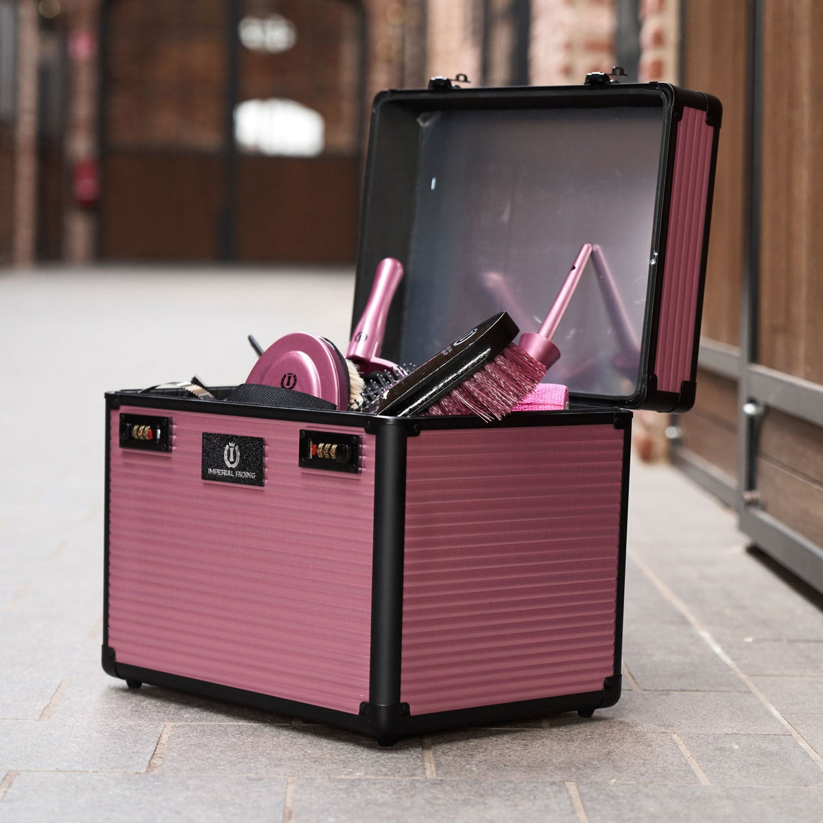 Mit Pferdepflege-Equipment gefüllte Imperial Riding Putzbox IRHShiny in Dark Mauve, stehend in der Stallgasse vor den Pferdeboxen. Die geöffnete Pferdeputzbox beinhaltet passende Bürsten, Striegel und Hufpinsel für die Fellpflege, optimal organisiert durch verstellbare und herausnehmbare Trennwände.