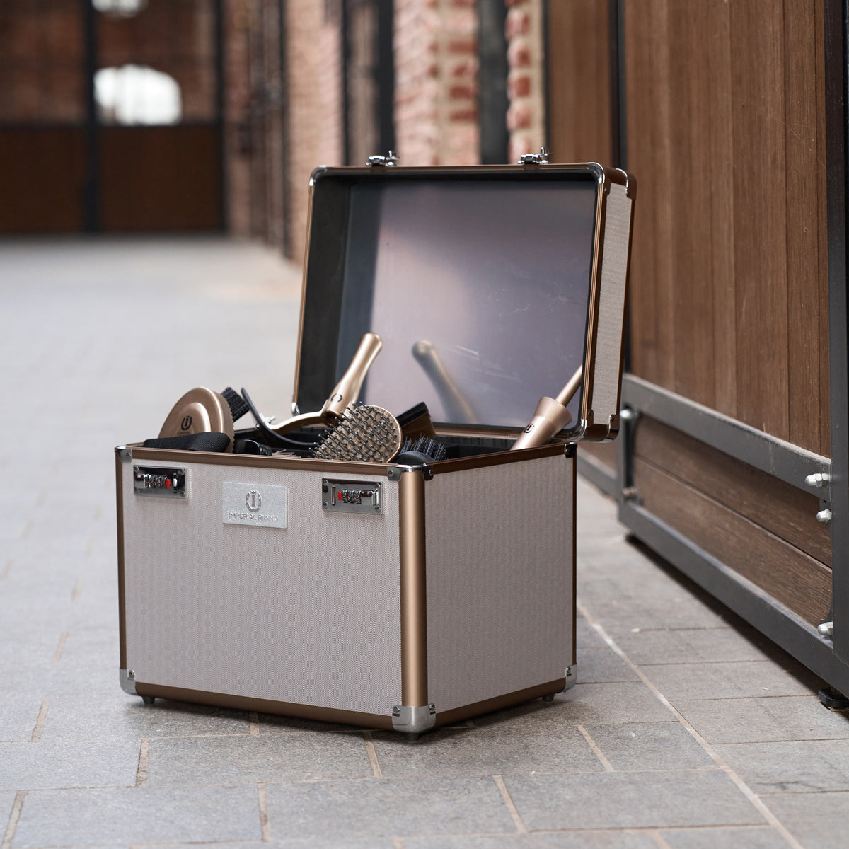 Befüllte Imperial Riding Putzbox IRHShiny Taupe Herringbone einsatzbereit in der Stallgasse. Die geöffnete Pferdepflegebox mit Spiegel, textilem Chevronmuster und roségoldenem Rahmen zeigt geordnetes Putzzeug wie Kardätschen, Striegel und Hufpinsel auf herausnehmbaren Trennwänden.