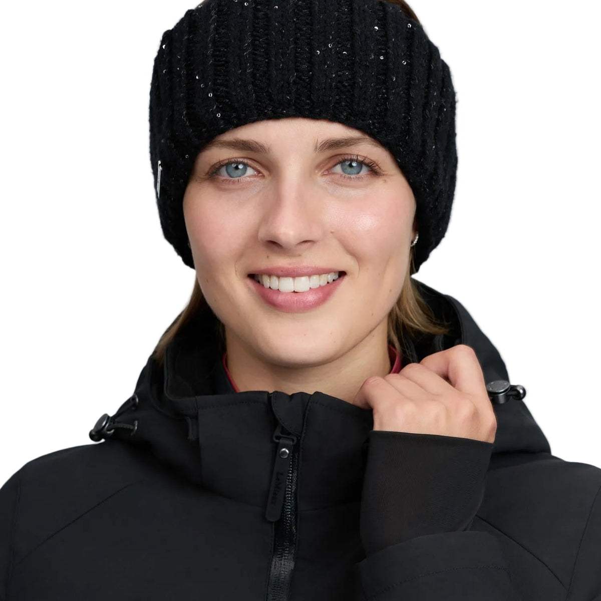 Nahaufnahme: Lächelnde Frau im warmen Winteroutfit mit hochgeschlossenem Kragen präsentiert das schwarze LeMieux Stirnband Bernie mit eingearbeiteten Pailletten. Der weiche Damen-Ohrenwärmer aus elastischem Rippstrick schützt Stirn und Ohren zuverlässig vor Kälte und stürmischem Wetter.