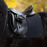 Nahaufnahme der schwarzen LeMieux Essence Dressurschabracke (Black) auf einem Rappen unter einem modernen Dressursattel. Das edle Chevron-Muster und der schützende PU-Leder-Bereich mit dem innovativen Gurtverriegelungssystem sind deutlich sichtbar. Diese Technik verhindert Verrutschen, Faltenbildung und Reibung des Sattelgurtes. Oben sichert die elastische D-Ring-Befestigung die Schabracke vor dem Hineinziehen in die Sattelkammer.