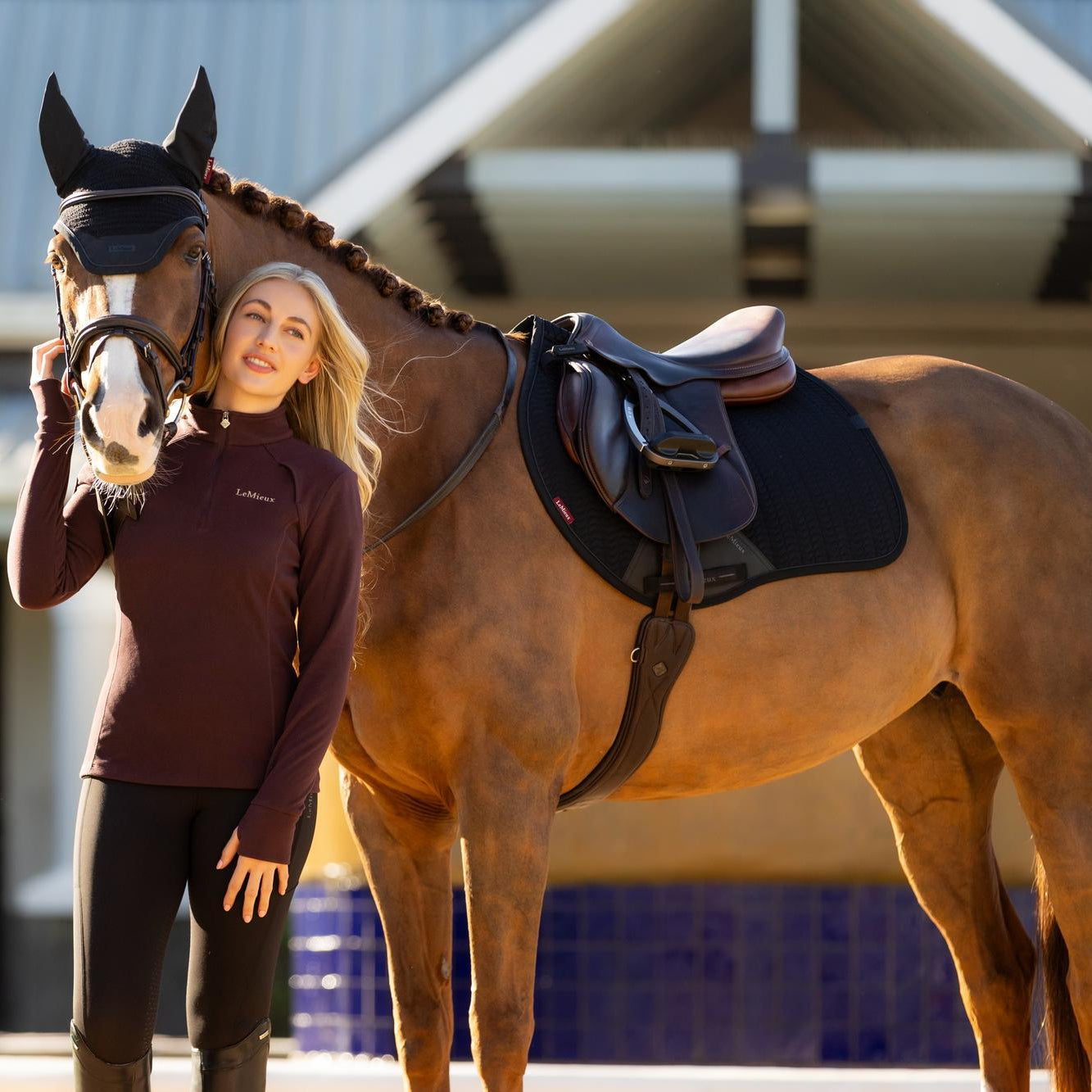Reiterin neben ihrem fuchsfarbenen Springpferd im kompletten LeMieux Essence Eurojump Set in Schwarz. Die anatomische Sattelunterlage mit Schwanenhals liegt perfekt unter dem Springsattel und bietet durch die spezielle Eurojump-Schnittform maximale Schulterfreiheit. Das atmungsaktive Softshell-Material der Essence Black Edition und das Self-Cool-Futter garantieren optimales Klimamanagement im täglichen Springtraining.