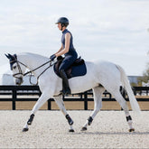 Ein weißer Schimmel im sommerlichen Training auf dem Reitplatz trägt das LeMieux Essence Set in Navy bei Sonnenschein. Während der Aufwärmphase unter der Reiterin sorgt das innovative Self-Cooling Material der Springschabracke für eine aktive Temperaturregulierung und verhindert Hitzestau. Die dunkelblaue Farbe bietet einen zeitlosen Kontrast zum hellen Fell und ist ideal für alle Pferdefarben geeignet.