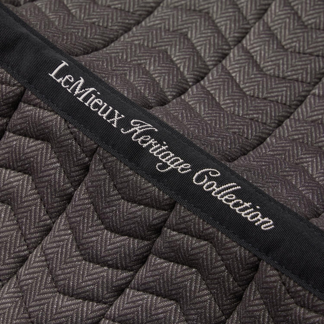 Detailaufnahme der LeMieux Heritage Dressurschabracke in Cocoa: Besticktes Topband der LeMieux Heritage Collection auf feiner Chevron-Steppung im braunen Fischgrätmuster. Das Topband verläuft mittig über die gesamte Rückenlinie. Die Bestickung dient der Kennzeichnung der Schabracke im Regal oder Sattelschrank.
