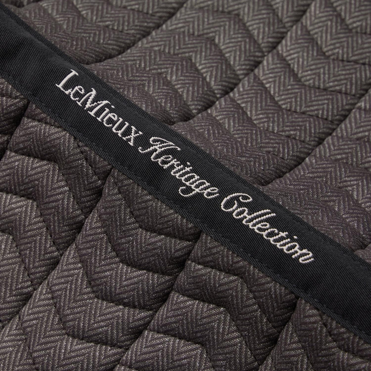 Detailaufnahme der LeMieux Heritage Dressurschabracke in Cocoa: Besticktes Topband der LeMieux Heritage Collection auf feiner Chevron-Steppung im braunen Fischgrätmuster. Das Topband verläuft mittig über die gesamte Rückenlinie. Die Bestickung dient der Kennzeichnung der Schabracke im Regal oder Sattelschrank.
