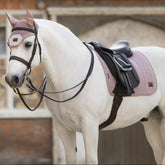 Dreiviertel-Ansicht eines Schimmels mit der LeMieux Heritage Herringbone Dressurschabracke im zarten Farbton Wild Rose (Altrosa) unter einem tiefen Dressursattel. Die Aufnahme zeigt die harmonische Abstimmung der gesteppten Schabracke mit anatomischem Schnitt und der passenden LeMieux Heritage Fliegenmütze als edles Set für den Dressursport.