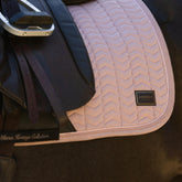 Nahaufnahme der LeMieux Heritage Dressurschabracke in Wild Rose (Altrosa) unter dem Sattelblatt eines Dressursattels. Deutlich sichtbar sind das edle LeMieux-Heritage-Logo-Badge, die feine Chevron-Steppung mit Fischgrätmuster sowie die zweifache Zierkordel aus gedrehter Kordel und Flechtkordel entlang der formstabilen Velours-Einfassung.