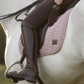 Detailaufnahme der LeMieux Heritage Dressurschabracke Herringbone in Wild Rose (Altrosa) im gerittenen Zustand auf einem Schimmel. Die Szene zeigt das elegante Reitoutfit mit braunen Lederreitstiefeln und Dressursattel sowie die feine Chevron-Steppung, die doppelte Zierkordel und das LeMieux-Heritage-Emblem unter dem Oberschenkel der Reiterin.