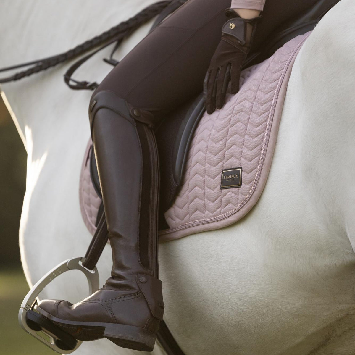 Detailaufnahme der LeMieux Heritage Dressurschabracke Herringbone in Wild Rose (Altrosa) im gerittenen Zustand auf einem Schimmel. Die Szene zeigt das elegante Reitoutfit mit braunen Lederreitstiefeln und Dressursattel sowie die feine Chevron-Steppung, die doppelte Zierkordel und das LeMieux-Heritage-Emblem unter dem Oberschenkel der Reiterin.