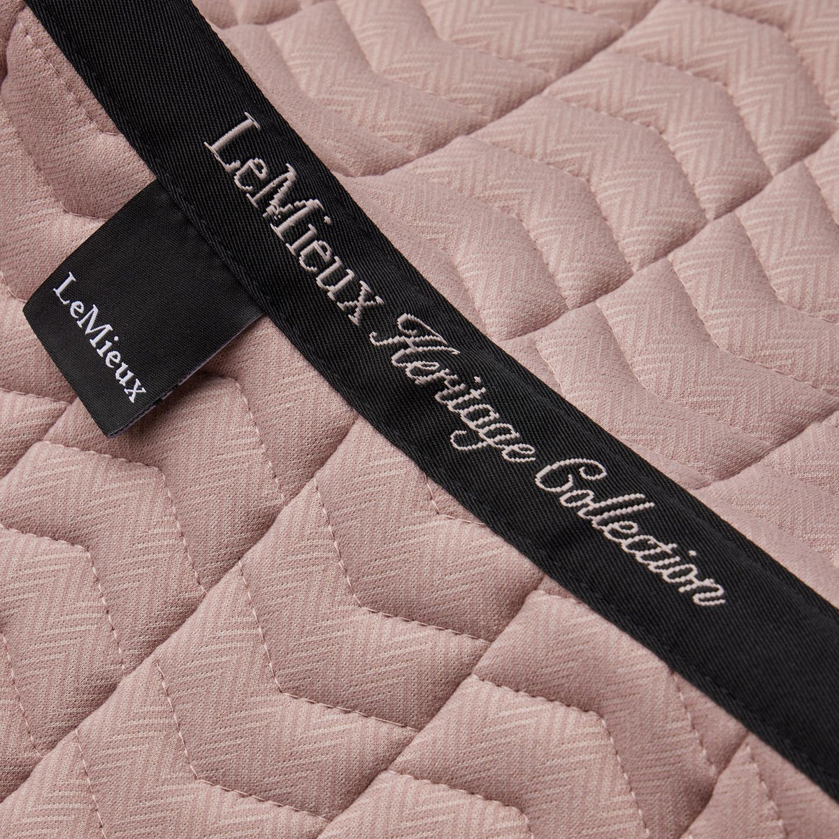 Detailaufnahme der LeMieux Heritage Dressurschabracke in Wild Rose (Altrosa) mit Fokus auf das edle Topband auf der Rückenlinie. Zu sehen sind die geschwungene weiße Stickerei „LeMieux Heritage Collection“, das seitliche LeMieux-Markenlabel und die feine Chevron-Steppung im Fischgrätmuster (Herringbone) auf dem pastellfarbenen Oberstoff.
