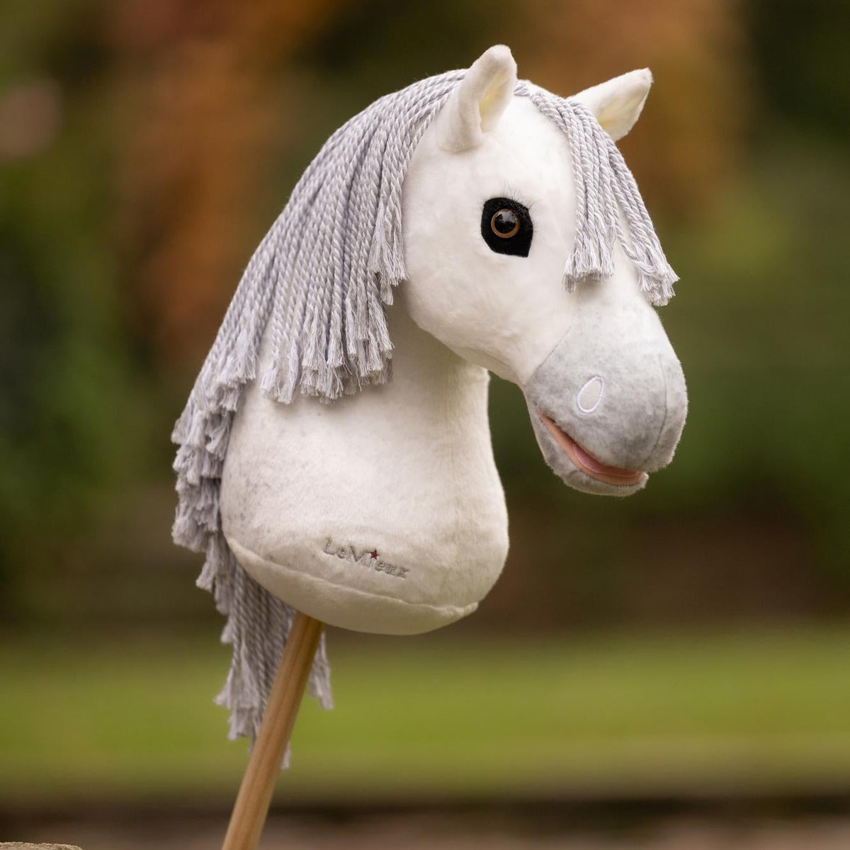 Das weiße LeMieux Hobby Horse Coco mit hellgrauer Mähne. Steckenpferd bei einem Ausritt in der Natur vor herbstlicher Kulisse, mit realistischer hellgrauer Mähne, rosa Maul und edler grauer LeMieux-Stickerei für authentisches Hobby Horsing und Turniertraining.