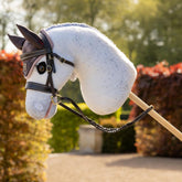 Das weiße LeMieux Hobby Horse Coco als Fliegenschimmel beim Training in englischer Parkkulisse, voll ausgestattet mit brauner Hunter Leder-Trense und passender Heritage Fliegenmütze in Cocoa mit Karomuster – bereit für schnelle Ritte und Sprünge.
