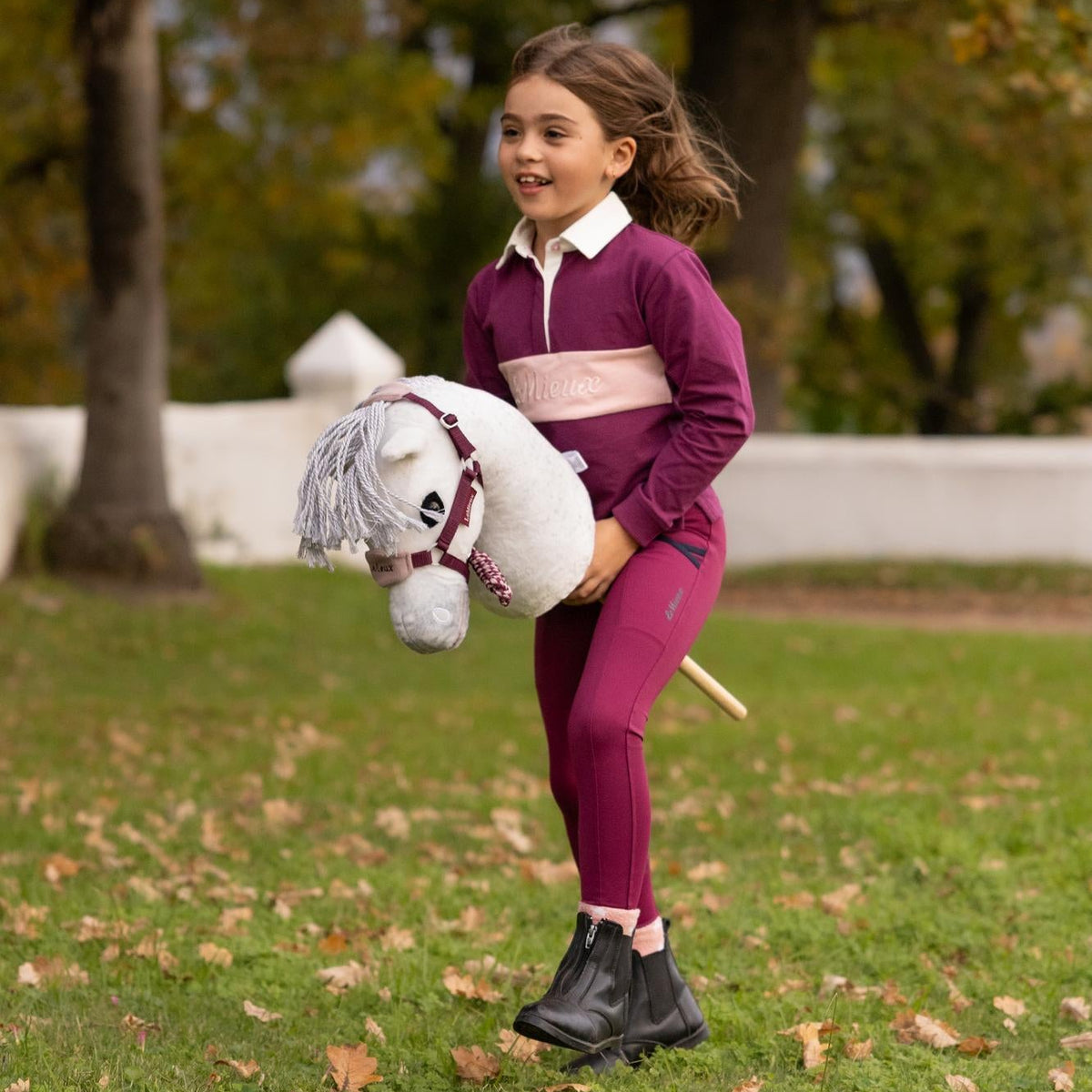 Ein Mädchen im Galopp auf dem weißen LeMieux Hobby Horse Coco Fliegenschimmel mit Vogue Halfter in der Farbe Blackberry, perfekt abgestimmt auf die violette Reitbekleidung beim dynamischen Hobby Horsing Training im herbstlichen Park.