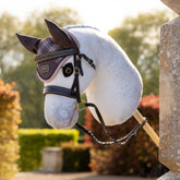 Detailaufnahme des LeMieux Hobby Horse Coco: Der weiße Schimmelkopf im Profil an einem Steinpfeiler, ausgestattet mit der braunen Leder-Trense Hunter und karierter Heritage Fliegenhaube in Cocoa für anspruchsvolle Hobby Horsing Turniere.