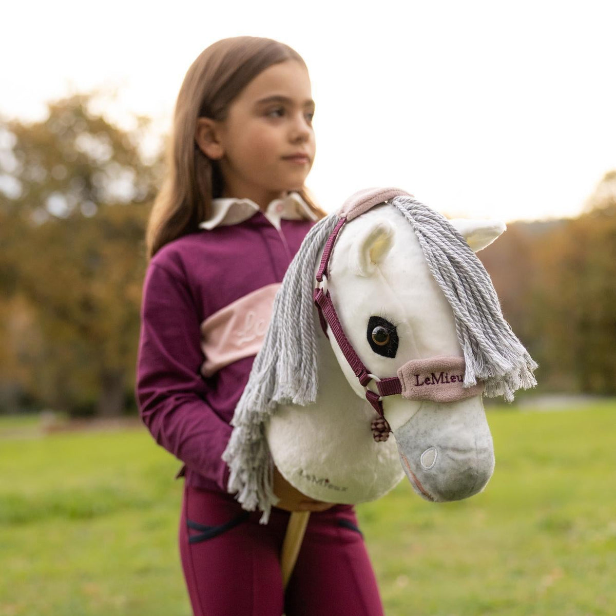 Porträt eines Mädchens mit dem LeMieux Hobby Horse Coco: Das weiße Schimmel-Steckenpferd trägt ein weich gepolstertes Vogue Fleece-Halfter in Blackberry mit Nasenschoner, farblich abgestimmt auf die Reitbekleidung für authentischen Reitspaß.