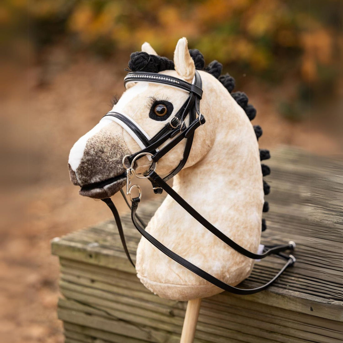Das LeMieux Steckenpferd Hobby Horse Dream, ausgestattet mit der passenden LeMieux Hobby Horse Dressurkandare aus Lack mit weißer Unterlegung. Das falbfarbene Hobby Horse steht draußen angelehnt in herbstlicher Umgebung . Für das Dressurreiten auf dem Turnier ist die schwarze Mähne kunstvoll geflochten und zu eleganten Rollzöpfen eingedreht. Ein realistisches Turnierpferd für Hobby Horsing.