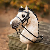 Das LeMieux Steckenpferd Hobby Horse Dream, ausgestattet mit der passenden LeMieux Hobby Horse Dressurkandare aus Lack mit weißer Unterlegung. Das falbfarbene Hobby Horse steht draußen angelehnt in herbstlicher Umgebung . Für das Dressurreiten auf dem Turnier ist die schwarze Mähne kunstvoll geflochten und zu eleganten Rollzöpfen eingedreht. Ein realistisches Turnierpferd für Hobby Horsing.