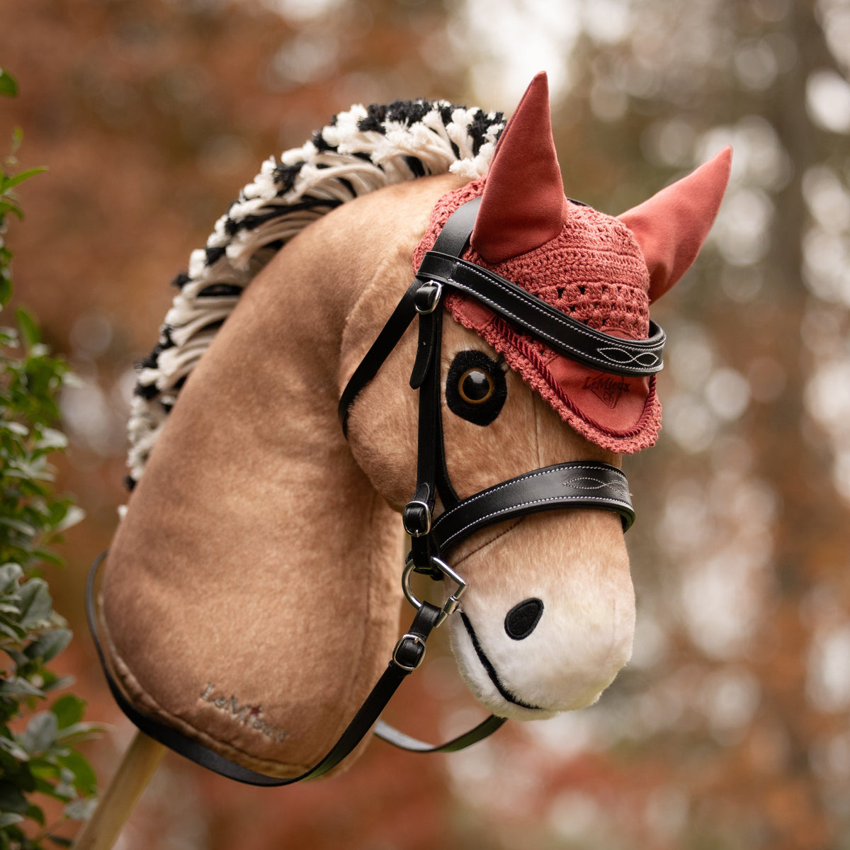 Die rosafarbene LeMieux Hobby Horse Fliegenhaube Spiced Earth auf dem Fjordpferd-Steckenpferd Oskar. Kombiniert mit der schwarzen Hobby Horse Trense Competition in herbstlicher Outdoor-Umgebung – perfektes Zubehör für Hobby Horsing Turniere, Training und Ausritte.