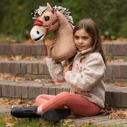 Mädchen sitzt im Freien mit dem LeMieux Hobby Horse Oskar (Fjordpferd Falbe) im Arm. Das Steckenpferd mit zweifarbiger Stehmähne trägt das passende gepolsterte LeMieux Hobby Horse Halfter mit Führstrick in Spiced Earth – realistisches Reitspielzeug für Kinder ab 3 Jahren.