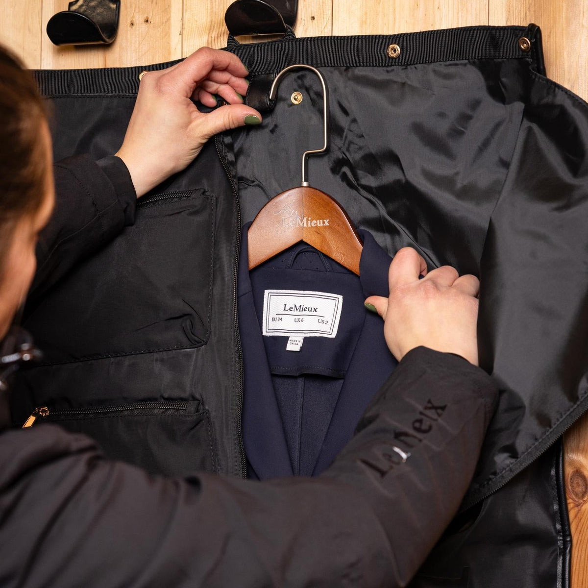 Detailansicht der praktischen Kleiderbügel-Schlaufe im schwarzen LeMieux Kleidersack: Ein dunkelblaues Turnierjacket hängt faltenfrei auf einem LeMieux Holzkleiderbügel im geöffneten Kleiderträger der Luxe Show Jacket Turniertasche.