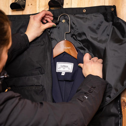 Detailansicht der praktischen Kleiderbügel-Schlaufe im schwarzen LeMieux Kleidersack: Ein dunkelblaues Turnierjacket hängt faltenfrei auf einem LeMieux Holzkleiderbügel im geöffneten Kleiderträger der Luxe Show Jacket Turniertasche.